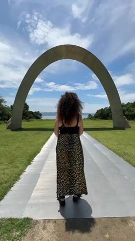 A person walks on a paved path towards a large concrete arch. The person is facing away from the camera, wearing a black tank top and a leopard-print skirt. The arch frames a view of the ocean and a lush green landscape under a partly cloudy sky. The path is flanked by green grass.