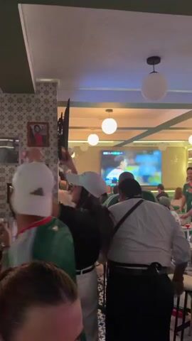 The video shows a group of people in what appears to be a bar or restaurant. Several individuals are wearing green jerseys with the Mexican flag colors, suggesting they are watching a sporting event, likely a soccer match. In the foreground, a person with their back to the camera is wearing a white baseball cap and a green jersey. To their left, another person is also wearing a white cap and a black shirt, with their hands on their head. In the background, a large screen displays a sports broadcast. The environment is dimly lit, with overhead lights providing illumination. There are decorative tiles on a pillar and framed pictures on the wall. Some people are seated, while others are standing and seem engaged with the screen. The overall atmosphere seems lively and focused on the event being broadcast.
