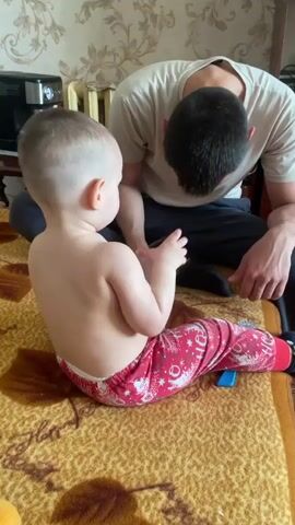 A toddler sits with his back to the camera on a brown and gold blanket wearing red pants with a white Christmas pattern, while an adult male sits behind the toddler on the same blanket with his legs crossed. The adult wears a light tan T-shirt and dark pants. The toddler holds up a head massager and puts it on the adult’s head. A coffee maker is on a table behind the adult and toddler. A patterned wallpaper is on the wall.