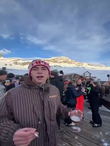 The video captures a lively après-ski scene in a mountainous area. Several people are visible, most holding drinks, suggesting a social gathering. The main subject, a young man wearing a striped jacket and a red and white headband, is holding a glass of beer. The backdrop showcases a snowy mountain range under a partially cloudy sky. The scene appears to be on a wooden deck or platform, with a building visible in the background, suggesting a ski resort or lodge. People are mingling and appear to be enjoying the atmosphere with music playing. The overall impression is that of a festive, outdoor celebration in a winter setting.