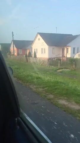 The video shows a view from inside a vehicle, looking out the side window. The scene outside is a rural landscape with houses and a road. The houses are colorful and appear to be simple dwellings. There is a wooden fence in front of the houses, and a grassy area between the road and the fence. The road is unpaved and appears to be made of gravel or dirt. The sky is clear and blue, with some wispy clouds. There are some trees and bushes visible in the distance. The video appears to have been recorded in the morning, as the sun is low in the sky and there is a slight mist. The vehicle is moving, as evidenced by the blur of the road and the passing houses. There are no people visible in the video. The overall impression is one of a peaceful and quiet rural setting.