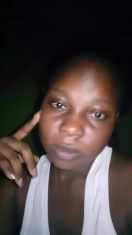The video shows a dark-skinned woman wearing a white tank top. She looks at the camera, sometimes glancing down. Her hand is raised near her face, with her fingers extended, seemingly touching her cheek. The background is dark, with indistinct green hues visible. The lighting is poor.