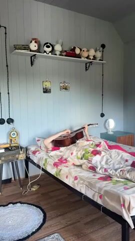 The video shows a person lying in bed under a floral comforter, playing an acoustic guitar. Their head is mostly obscured by the guitar and blankets. The room has light blue walls with white paneling on the ceiling. A white shelf above the bed holds several stuffed animals and books. Two framed pictures hang on the wall. To the left of the bed is a nightstand with a vintage clock and some items on top. A round rug is on the wooden floor. A dresser with a mirror is visible on the right side of the room. The lighting suggests daytime, with soft light entering the room.