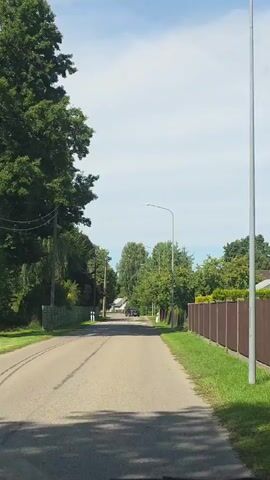The video shows a view from a vehicle driving down a paved road lined with trees and fences on both sides. There is a car ahead on the road, and streetlights are visible. The sky is blue with some clouds.
