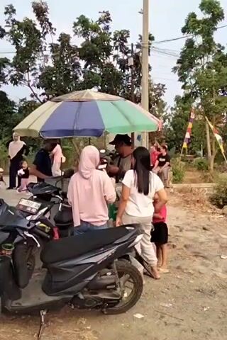 A group of people are gathered under a multi-colored umbrella. Two motorcycles are parked in the foreground. In the background, there are trees and a pole with wires. Some people are standing, while others appear to be interacting with a vendor under the umbrella. One person is holding a white cup. The ground is dusty and uneven. The overall impression is a casual outdoor gathering, possibly a food stall or marketplace.