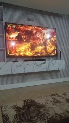 The video shows a living room with a large flat-screen TV mounted on the wall. A fighting video game is playing on the TV. A long, low white cabinet with a marble pattern sits under the TV. The wall is covered in a vertically striped material. There is a beige rug on the floor with a dark brown swirling pattern. A white shelf unit is visible on the left side of the frame. Someone is playing the game, but they are not visible in the video. The audio contains sound effects from the game and someone speaking in another language.