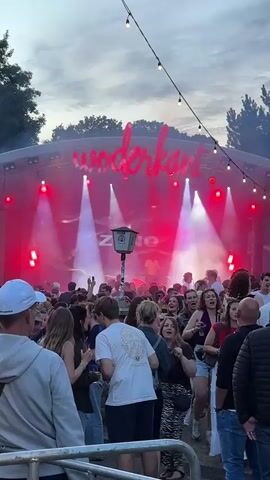 A crowd of people is gathered in front of a stage at an outdoor event. The stage has a large banner that says "woderkant" in red, illuminated letters. Red and white spotlights are shining on the stage and the crowd. Some people are dancing, and others are talking and socializing. The sky is visible above the stage, with some clouds and string lights overhead. In the foreground, several people are facing away from the camera, looking towards the stage. The overall atmosphere appears to be festive and energetic.