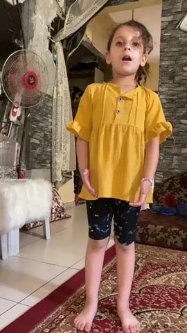 This is a vertical, eye-level video of a young girl in Khan Yunis, Palestinian Territories. The girl, with brown curly hair, is standing on a patterned rug in what appears to be a living room. She is wearing a yellow, short-sleeved tunic with ruffles and buttons, and black shorts with a colorful pattern. She has bracelets on her left wrist. Her mouth is open as if she is singing or speaking, and her hands are moving expressively, at times held together, at others extended outwards, and once placed on her hips. 

The setting is modestly decorated, with a brick-patterned wall behind the girl, and a large, red-bladed fan to her left. A white stool with a fluffy cover is also visible. A patterned sofa is partially visible on the right. The lighting suggests it is daytime, and the atmosphere is casual and domestic. The girl's facial expression and energetic gestures convey a sense of joy and engagement in whatever she is doing. There is no visible text in the video.