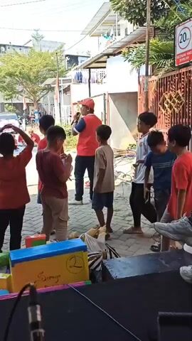 A group of children and adults are gathered outdoors, likely for an event or celebration. The scene appears to be in a residential area, with houses and trees visible in the background. Several children, mostly wearing red, are standing in a semi-circle, facing a man who is holding a microphone and speaking. One child in the center is holding what appears to be a small, wrapped object. Another adult, possibly a woman in a hijab, is also present and interacting with the children. Colorful boxes are stacked on the ground in the foreground, suggesting a game or activity. The lighting indicates it is daytime, with bright sunlight. There are also some vehicles and other people visible in the background, suggesting a community gathering.