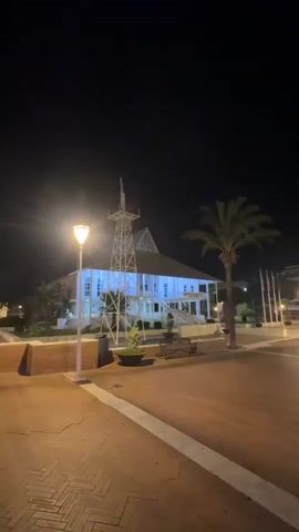 A night scene shows a building illuminated with blue lights, with a tower structure in front of it. A street lamp is on the left side of the frame, casting light on a brick pathway. A palm tree stands to the right of the building, and a line of flags is visible behind it. The street in front of the building is lit by streetlights, and a car is driving on the right side of the street. The sky is dark, indicating it is nighttime.