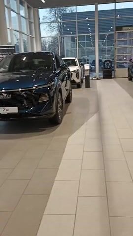 The video shows the interior of a car dealership. Several cars are on display. The nearest car is a dark blue Haval F7x. Behind it is a white car and another blue car. The dealership has large windows. The floor is made of large, light-colored tiles.