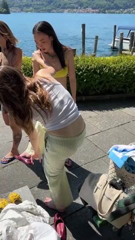 Three women are by a lake. One woman wears a sundress and sunglasses and adjusts her sunglasses. Another woman wearing a white top and striped pants bends over, handling maroon shoes. A third woman in a yellow top stands behind her. Bags and shoes are on the ground. The camera pans to show a view of the lake, a building on an island, and mountains in the background. A woman wearing a red plaid hat smiles at the camera. There are people and boats in the background. The day is sunny.