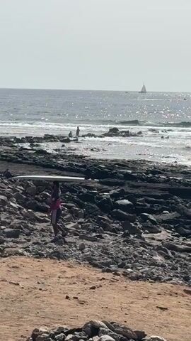 A person carrying a surfboard walks along a rocky beach towards the ocean. The ocean is calm with small waves and a sailboat is visible in the distance. The sky is overcast.