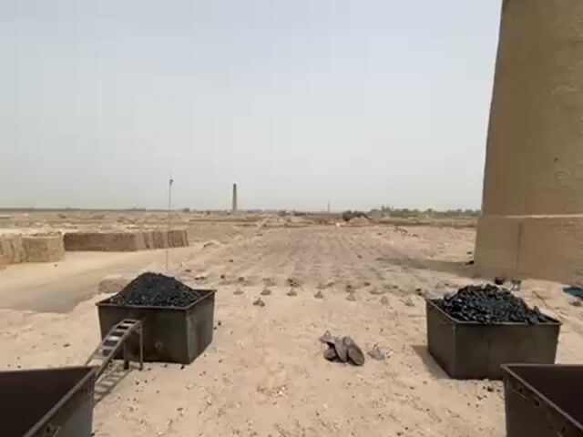 The video shows a brickyard. In the foreground, there are three black plastic bins, two filled with coal. A small metal ramp leads to one of the coal-filled bins. There are various metal objects, including what appears to be footwear, on the ground between the bins. A tall, cylindrical brick chimney stands prominently behind the bins. The brickyard stretches into the distance, with rows of bricks laid out on the ground and another chimney visible in the background. The sky is overcast.