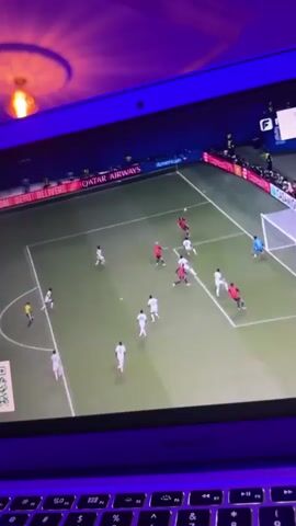 A close-up shot shows a laptop screen displaying a soccer game. The screen is illuminated by blue light, casting a purple glow on the laptop's keyboard and the wall behind it. The game shows players in red and white uniforms on a green field, with a goal at the right side of the screen. One player in red kicks the ball towards the goal. The camera is positioned above and slightly to the left of the laptop.