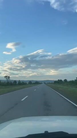 This media file is a first-person perspective video of driving on a straight, paved road. The road has white lane markings and is bordered by grassy fields and scattered trees. In the distance, rolling hills create a layered horizon. The sky is a vibrant blue with scattered, wispy white clouds, suggesting a pleasant day, possibly late afternoon or early evening due to the soft lighting. The overall scene evokes a sense of journey and open space. There are no people or specific objects visible other than the road, vegetation, and the part of the vehicle from which the video is being recorded. The setting appears to be rural, consistent with the provided location context of Khumalag, Russia. The mood is tranquil and expansive.