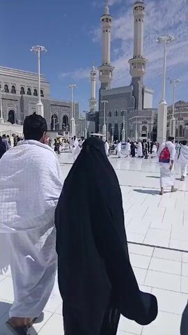 The video shows a couple walking across a white tiled plaza toward the Masjid al-Haram in Mecca. The man is wearing a white Ihram, and the woman is wearing a black niqab. They are surrounded by other pilgrims. The building in the background has tall minarets, and the sky is blue.