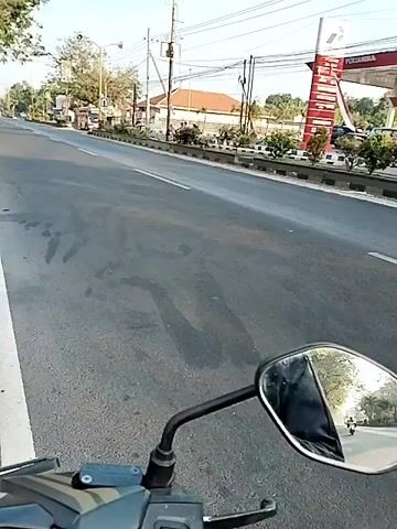 The video shows a view from a motorcycle as it travels on a paved road. In the foreground, a motorcycle's rearview mirror reflects the rider's hand and arm, which are clad in dark clothing. The mirror also shows a glimpse of the road behind the motorcycle, with another vehicle in the distance. The background features a gas station with red and white signage, likely indicating a Pertamina station. Several vehicles are visible at the gas station, including cars and motorcycles. A bus is seen driving past the gas station. The road is lined with a concrete barrier and small trees or bushes. Power lines are visible overhead. The sky is clear and bright, suggesting it is daytime. The overall scene depicts a typical roadside environment with traffic and commercial establishments.