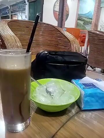 A coffee drink, a bowl of ice, a pack of tissues, and a black bag sit on a wooden table in a public space. The coffee drink is in a tall, clear glass with a black straw. The bowl is light green and filled with small, irregular ice cubes with a spoon inside. The tissues are in a blue box. A black bag sits behind the bowl of ice. The background shows rattan furniture, paintings on the walls, and the exterior of a building with various signs and posters.