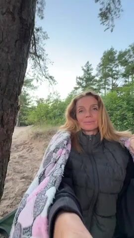 This media file depicts a tranquil scene on a sandy beach in Peski, Russia. The primary subject is a woman with blonde hair, who appears to be relaxing by a campfire. She is wrapped in a blanket adorned with a pink and white patterned design and wearing a dark puffer jacket. She is looking directly at the camera with a gentle smile, suggesting a feeling of contentment.

The setting is a beach with sand dunes and sparse vegetation. A campfire is actively burning in a metal brazier, with smoke rising into the air. Logs are arranged around the brazier, indicating preparation for or continuation of a fire. A green camping chair is positioned to the left of the campfire, adding to the impression of a relaxed outdoor setting. In the background, pine trees frame the horizon, and beyond them, a calm body of water, likely the sea or a large lake, is visible. The sky exhibits the soft hues of either dawn or dusk, with pastel colors painting the horizon, suggesting a peaceful time of day. The overall atmosphere is serene and suggests a moment of quiet enjoyment in nature.