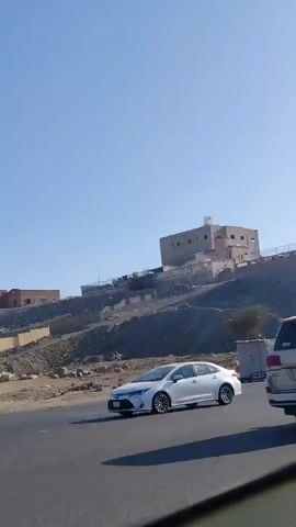 The video shows a street scene with a building on a hill in the background, filmed from inside a moving car. The sky is clear and blue. Several cars pass in the street. A person walks on the side of the road at the beginning of the video. There are also some small buildings alongside the road, one of which has writing on it.