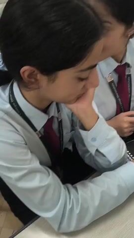 The video shows three young women, likely students, gathered around a table. They are all wearing light blue collared shirts and maroon ties, suggesting a school uniform. One woman in the foreground is leaning forward, resting her chin on her hand, and looking down at a notebook. She appears to be engaged in a conversation or task. To her right, another woman is looking up and smiling, holding a blue pen and pointing at something on a notebook. A third woman is visible in the background, also looking down at a notebook and holding a pen. The environment appears to be a classroom or study area, with a wooden table and chairs. There are notebooks, pens, and a student ID card visible on the table. The lighting is neutral, and the focus is on the individuals and their interactions.