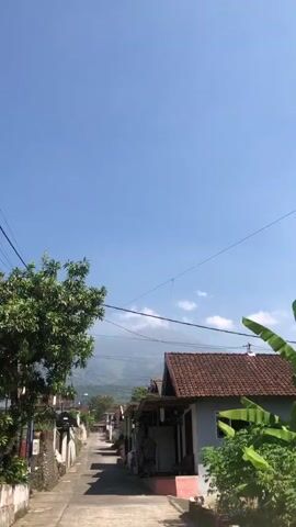 The video shows a narrow, paved road lined with houses on both sides. The sky is clear and blue. There are trees and power lines visible. The houses have red tile roofs and some have awnings or covered porches. In the distance, there are mountains. The road appears to be in a residential area.