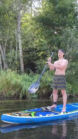 This short video captures a young man paddleboarding on a calm body of water, likely a river or lake, surrounded by lush greenery. He is shirtless, wearing checkered shorts and a baseball cap, and is holding a paddle. The man appears focused as he navigates the water, his posture suggesting a relaxed yet purposeful stance.

The scene is set in a natural environment with dense trees and tall grasses lining the bank. The sunlight filtering through the leaves suggests it is daytime, possibly late afternoon, given the soft lighting. The water is still, reflecting the surrounding vegetation and the blue sky.

The paddleboard itself is blue with colorful accents and text visible on its side. Part of the text appears to read "Feath-R-Lite," possibly indicating the brand of the paddleboard. The man has an ankle leash attached to his leg and the board, a common safety feature for paddleboarding.

There are no explicit emotional cues, but the overall atmosphere is peaceful and serene, characteristic of outdoor recreational activities in nature. The location is identified as Starosiverskaya, Russia, which places this scene within a specific geographical context of forested landscapes and waterways typical of the region. The video is short, focusing on the simple act of paddleboarding in a tranquil setting.