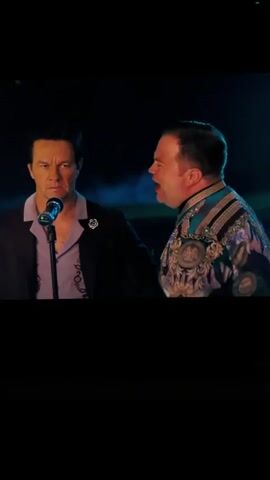 Two men are on a stage. The man on the left is wearing a black suit jacket over a purple shirt. He is standing at a microphone and appears to be singing or speaking into it. His face is contorted in an angry expression. The man on the right is wearing an elaborate, sequined costume with gold and blue accents. He is standing to the left of the first man and his mouth is wide open, as if he is shouting or singing loudly. The background is dark with some hazy blue and white lighting, suggesting it is nighttime or a dimly lit indoor venue. The camera is focused on the two men.
