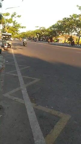 The video captures an outdoor scene on a street with trees lining the right side of the road. The road appears to be paved with asphalt and has lane markings. In the foreground, a yellow and white striped marking is visible on the side of the road. Several motorcycles are parked on the left side of the road. Traffic is present on the road, with at least two motorcycles and one car visible in the distance. The sun is shining, casting shadows on the road, suggesting it is daytime. The overall environment appears to be a street in a town or city.