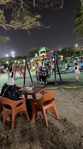 The video captures a nighttime scene at a park or playground. In the foreground, two orange chairs and a small table are set up on a sandy surface. Beyond this, a playground area is visible with various equipment, including swings and slides. Several children and adults are present in the playground area, with some children on the swings and others near the slides. In the background, trees with foliage are illuminated by artificial lighting. Further in the distance, there appear to be tables and chairs set up, possibly for an outdoor seating area, and some buildings are visible. The overall atmosphere suggests a family-friendly, recreational environment.