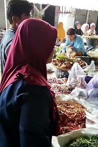 The video shows a bustling outdoor market scene. In the foreground, a person wearing a burgundy hijab is partially visible, facing away from the camera and looking towards a market stall. The stall is laden with fresh produce, including large piles of red chilies, onions, and green vegetables. Behind the foreground figure, several people are engaged in commerce. A woman in a blue shirt and a brown cap is sorting vegetables. Another woman in a patterned shirt and black hijab is preparing produce, placing items into a plastic bag. Other individuals, mostly women wearing hijabs in various colors, are seen browsing or attending to their stalls in the background. The market appears to be covered by a light-colored awning or roof structure. The overall impression is one of a vibrant, active marketplace.