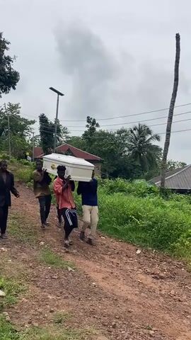 A group of four men are carrying a white coffin on a dirt path. The man in the front is wearing a black suit and black pants. Behind him, three other men are carrying the coffin. The man in the middle is wearing a red shirt and black shorts with white stripes. The man to his left is wearing a green t-shirt and black pants. The man to his right is wearing a blue t-shirt and khaki pants. The men are walking uphill, and the coffin is angled slightly upwards. The environment is rural, with lush green vegetation on the sides of the path. There are houses and trees in the background, and the sky is cloudy.