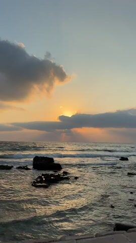 This is a static, eye-level shot of a sunset over the ocean. The sun is partially obscured by clouds, casting rays of light onto the water. The ocean is calm with small waves. There are rocks in the foreground, some of which are submerged in the water. The sky is a gradient of orange, yellow, and blue. There are no people or animals visible in the video.