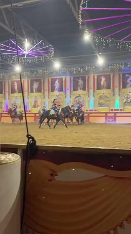 A horseback riding performance is taking place in a dimly lit indoor arena. The arena floor is covered in sand. In the background, a decorative backdrop resembling ancient Roman architecture with statues and murals is illuminated by colorful lights. Several riders, dressed in colorful costumes, are on horseback in the arena. Some horses are in motion, while others appear to be standing still. The riders seem to be in formation or performing some coordinated maneuvers. The overall atmosphere suggests a theatrical or circus-like event.