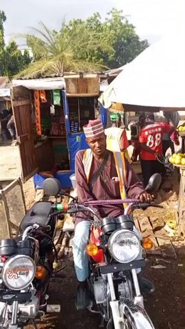 The video shows a street scene with several motorcycles parked. In the foreground, a person wearing a blue traditional outfit and a cap is looking at their phone while standing next to a row of motorcycles. To the right of the person, another motorcycle is visible. In the background, a fruit stand is set up under a large umbrella, with various fruits displayed on tables. Several people are present at the fruit stand, including one wearing a yellow vest and another in a red shirt. A person is also seen sitting on a motorcycle in the left foreground of the video and gesturing with their hands. The ground appears to be dirt or gravel, and there are some puddles of water, suggesting it may have recently rained. The overall environment appears to be a bustling outdoor market or street in a town or city.