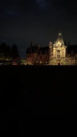 The video shows a large, stately building illuminated at night. The building has multiple stories and a prominent tower. The lights are on inside and outside the building, casting a warm glow. The sky is dark and cloudy, with some faint lightning visible in the distance. There is a large, dark area in the foreground, possibly a lawn or field. Trees are visible on the left side of the building. The overall impression is one of grandeur and mystery.