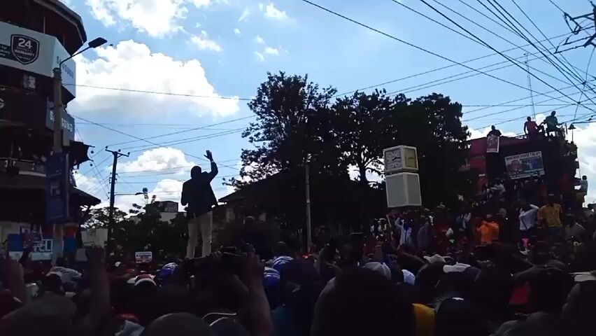 Here is a description of the video:

The video shows a dense crowd of people gathered in an urban environment, possibly for a political rally or public demonstration. The crowd is dense with people raising their hands and holding up phones, seemingly to capture the event.  

Standing above the crowd, a man is visible on top of a vehicle or structure, giving a speech using a microphone. A  speaker system can be seen positioned nearby, likely used for broadcasting the speech. Banners or posters with political messages are visible in the background, indicating the nature of the gathering. The sky above is partly cloudy, and there are buildings and utility poles with wires surrounding the scene.