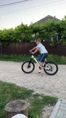 This video captures a young boy riding a white bicycle down a gravel path in a rural setting. He is wearing a light blue t-shirt and dark blue shorts. The boy is positioned in the center of the frame, with the bicycle's front wheel turned slightly. The path is flanked by lush green grass and some scattered rocks. In the background, there's a wooden fence and dense green foliage, with houses and trees visible further down the road. Power lines and utility poles are also prominent features in the scene.

The time of day appears to be late afternoon or early evening, as indicated by the soft, warm lighting and the dusky sky with wispy clouds, suggesting a sunset. The weather seems pleasant and calm. The location is implied to be a village or countryside, likely in Pîrlița, Moldova, given the architectural style of the houses and the overall rural ambiance.

The boy's posture suggests he is actively engaged in riding, perhaps learning or practicing. There are no visible signs of text within the video. The overall mood is one of childhood freedom and simple outdoor activity. A blurred figure, possibly an adult filming, is visible in the foreground of some frames, adding a personal perspective to the recording. A cat is also visible in the grass in the foreground of one frame.