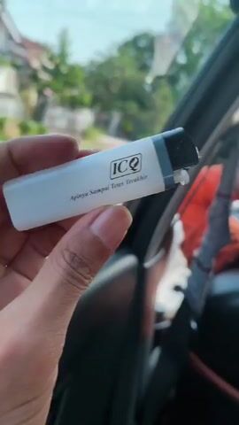 A hand holds a white cigarette lighter with the ICQ logo and text that reads "Apinya Sampai Tetes Terakhir" (Its fire until the last drop). The lighter is in focus, while the background, seen through a car window, is blurred and shows a residential area with trees and buildings. The video is a close-up shot of the lighter, with a slight shake that suggests movement.