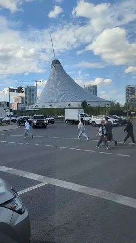 The video shows a street scene with a large, cone-shaped building in the background. Several cars are driving on the road, and pedestrians are crossing the street. The sky is blue with some clouds. The camera is positioned low, looking up at the scene. In the foreground, part of a silver car is visible. A traffic light is visible on the left side of the frame. People are walking across the crosswalk. A person on a bicycle with a yellow bag on their back is also crossing the street.
