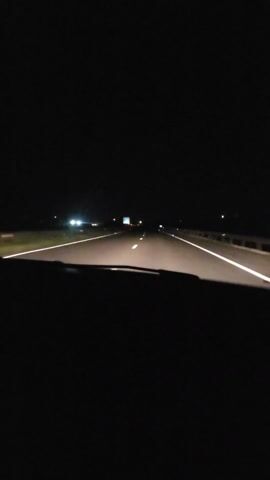 A car drives down an empty highway at night. The only light comes from the headlights, illuminating the white lane markers. Distant headlights of other vehicles can be seen ahead. The road is flat and straight, with some vegetation visible on the sides.