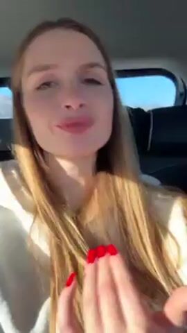 The video shows a woman with long blonde hair sitting in the back seat of a car. She is wearing a white hoodie and has red nail polish. The car appears to be moving, as scenery can be seen through the window. Loud music is playing in the background. The woman is smiling and looks directly at the camera.