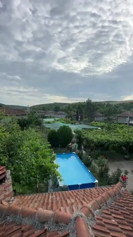 This aerial view captures a serene village scene in Tsarevets, Bulgaria. The foreground is dominated by the weathered, terracotta tiles of a rooftop, suggesting a traditional architectural style. Beyond this, a vibrant blue swimming pool stands out amidst lush greenery, a clear indicator of leisure and relaxation.

The middle ground reveals a collection of houses nestled amongst trees and neatly cultivated gardens. Some structures appear to have pergolas or vine-covered awnings, typical of rural European settings. A prominent element is a series of green-covered structures, possibly greenhouses or shaded cultivation areas, suggesting agricultural activity within the village. The presence of a Bulgarian flag on a flagpole indicates a sense of national identity.

The background is characterized by rolling hills covered in dense vegetation, hinting at a temperate climate and rural landscape. The sky is a dramatic canvas of clouds, with patches of sunlight breaking through, suggesting either early morning or late afternoon. The lighting and cloud formations create a slightly overcast but picturesque atmosphere, evoking a sense of calm and natural beauty. No people are visible in the frame, emphasizing the peaceful, uninhabited quality of the scene. The overall impression is one of a quiet, idyllic rural community enjoying a summer day.