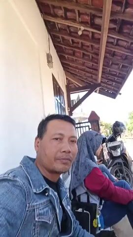 A man in a denim jacket is sitting close to the camera, looking at it. Behind him, two women are sitting on a porch. The woman in the foreground is wearing a maroon top and blue jeans. The woman behind her is wearing a patterned headscarf and blue jeans. A motorcycle is parked behind the women. The porch has a tiled roof and white walls. There is a door on the left side of the frame. The ground is covered in gravel. The scene is lit by natural sunlight.