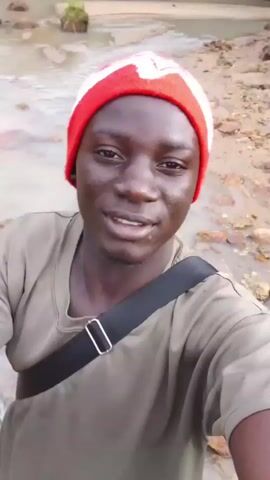 A close-up, selfie-style video shows a young Black person wearing a red and white beanie and a green t-shirt, with a black strap across their chest. They are standing in shallow, muddy water with rocks visible. The person smiles and looks at the camera. The background consists of muddy terrain and some greenery. The video is short and appears to be taken outdoors.