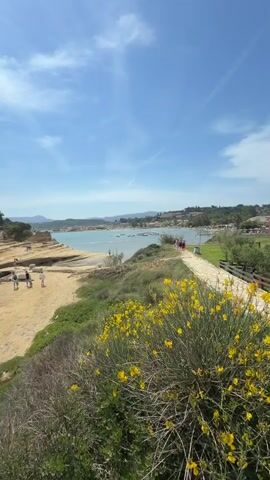 The video shows a coastal landscape on a sunny day with a blue sky and some clouds. In the foreground, there is a rocky cliff with green vegetation and yellow flowers. Below the cliff is a sandy beach with several people walking and standing. To the right, a paved path runs alongside the beach, with some trees and more people visible. The background features a calm turquoise sea with a few boats and a distant town with buildings. The camera pans slightly to the left, offering a wider view of the beach and the sea.
