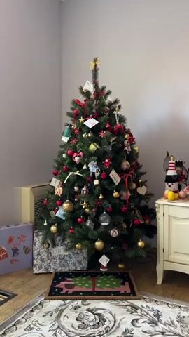 The video shows a decorated Christmas tree in a room. The tree is adorned with red and gold ornaments, ribbons, and small figures. Some of the ornaments appear to be handcrafted. There are also greeting cards hanging on the tree. To the left of the tree, there is a beige air conditioner and two decorative boxes. One box is purple with a gingerbread man design, and the other is grey and white with a pattern. In front of the tree is a small rug with a Christmas tree design. To the right of the tree, there is a white cabinet with decorative items on top, including a lighthouse-shaped object and some fruit. A patterned rug is visible on the floor in the foreground. The walls of the room are painted a light grey. The video is a close-up shot of the Christmas tree, with the camera slowly zooming in and panning slightly to the right.