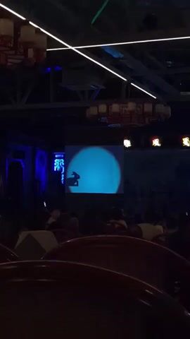 A wide shot shows a dimly lit room with a projector screen displaying a silhouette of a rabbit against a blue circle. The screen is positioned above a seated audience, whose backs are partially visible. The room has a high ceiling with exposed beams and decorative lighting fixtures. Tables with white cloths are visible in the foreground. The overall ambiance suggests an indoor event or performance.