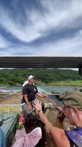 A man in a black t-shirt, white cap, and grey shorts is steering a long, narrow boat with a wooden oar. He is smiling and looking to his left. Several passengers are seated in the boat behind him. The boat is moving down a river with green, tree-covered hills in the background. The sky is partly cloudy. The boat appears to be passing under a bridge or some kind of overhead structure. There is writing in Japanese on the side of the boat.